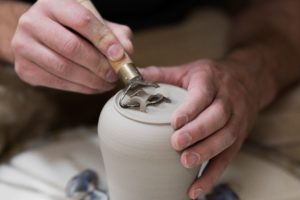 Zwei kaukasisch aussehende Hände arbeiten an einer Tonvase. Die eine Hand hält die Vase, die andere hält ein Schlaufenwerkzeug, mit dem sie vorsichtig eine Rille in den Vasenboden herauskratzt.